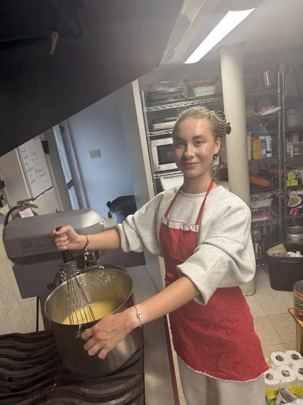 Anastasiia Stepanenko in a red apron, whisking batter in a large steel pot in a commercial kitchen.